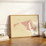 Framed Maryland county map leaning against a wall in a bright interior with warm wood flooring and minimal décor.