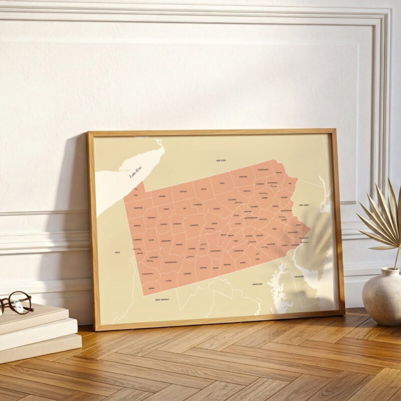 Framed Pennsylvania map print leaning against a white wall on herringbone wood flooring, styled with warm light and minimal accents.