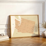 A framed Washington state map resting against a white wall on a wooden floor, styled with warm light and subtle decorative accents